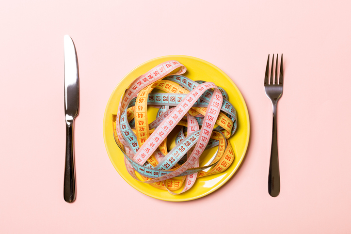 Top view of colorful measuring tapes on plate in the form of spaghetti with knife and fork on pink background. Weight loss and diet concept